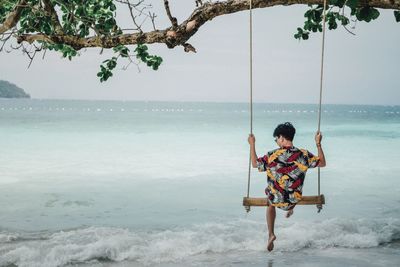 Rear view of man swinging at beach against sky