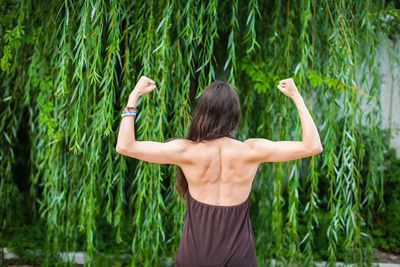 Rear view of woman flexing muscles while standing against plants