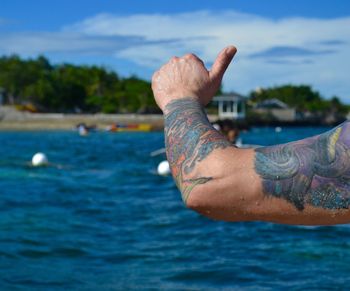 Cropped image of man with tattooed hand gesturing thumbs up sign at beach