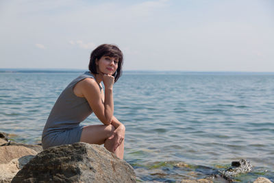 A happy adult woman is sitting on a rock on the riverbank sunbathing on a sunny day.