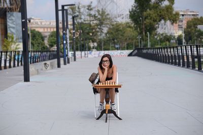 Portrait of woman sitting with chess board on chair at walkway