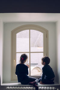 Rear view of brother and sister sitting by window at home