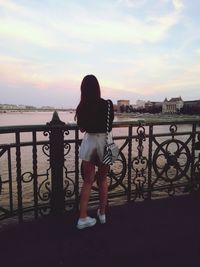 Rear view of woman standing by railing against sky during sunset