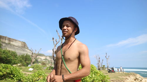 Shirtless young man standing at beach against sky