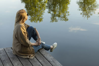 Young pretty blonde woman sit on wooden dock enjoying lake view in summer time on sunset. rest and