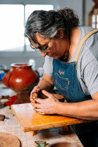 Side view of mature focused female craftswoman ceramist in apron shaping piece of clay pot in hand in workshop table