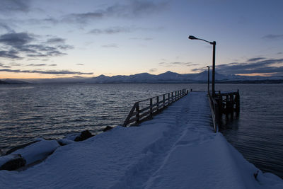 Snow covered pier by lake against sky during sunset