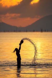 Side view of silhouette woman throwing water in lake against mountains during sunset