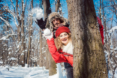 Portrait of happy friends throwing snow while standing behind tree trunk