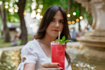 Beautiful young woman drinking strawberry mojito cocktail in park in city