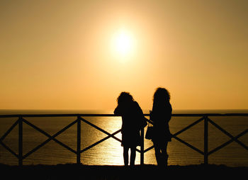 Silhouette women standing on railing against sea during sunset