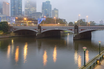 Bridge over river by illuminated buildings against sky at night