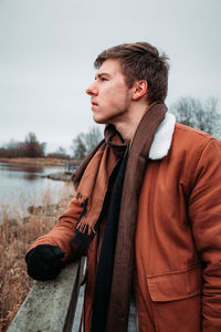 Young man looking away while standing by lake against sky