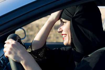 Side view of smiling woman wearing hijab driving car seen through window