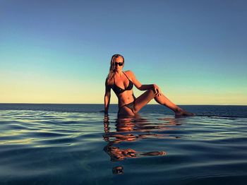 Young woman in bikini sitting on edge of infinity pool against clear sky