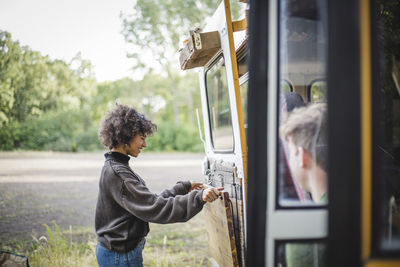 Side view of smiling woman adjusting wood on caravan door during camping in forest
