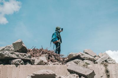 Low angle view of man photographing with mobile phone while standing on old ruin against blue sky during sunny day