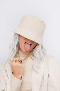 Close-up of woman wearing hat sticking out tongue against white background