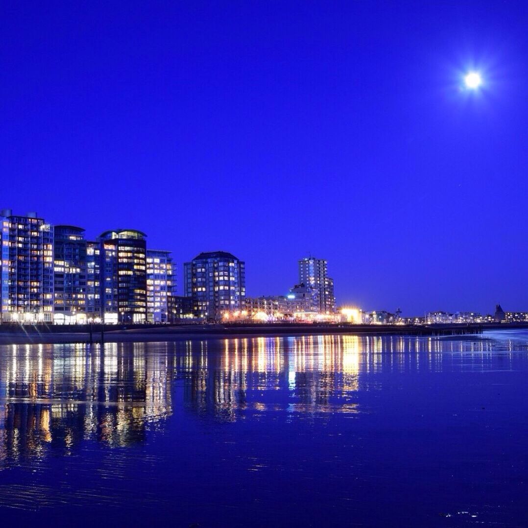 illuminated, water, night, waterfront, blue, reflection, building exterior, architecture, built structure, city, clear sky, copy space, cityscape, sea, river, sky, moon, outdoors, no people, mid distance