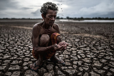 Shirtless man holding falling water while crouching on cracked land against sky