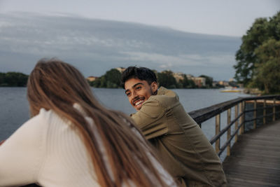 Side view of happy young man talking with female friend while leaning on railing at pier