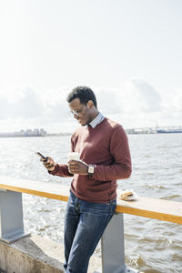 Young man standing at the sea, holding coffee, using smartphone