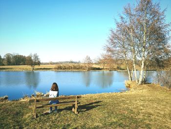 Rear view of teenage girl looking at sea while sitting on bench against clear sky
