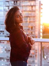 Portrait of young woman standing by railing in balcony during sunset