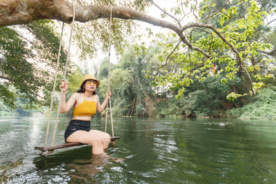 Young woman sitting on rope swing in river at forest