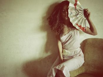 Young woman holding hand fan while sitting on sofa against wall