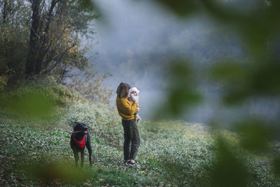 A woman is holding a baby and looking at a dog near a river