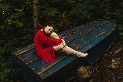 Young woman sitting on old boat against trees in forest