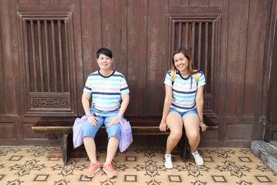 Portrait of smiling friends sitting on bench against wall