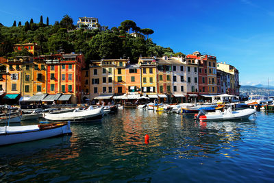 Boats moored in sea by buildings against blue sky