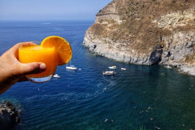 Cropped hand holding orange juice in glass against sea 