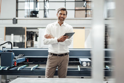 Smiling mature businessman holding digital tablet while standing against machinery in factory