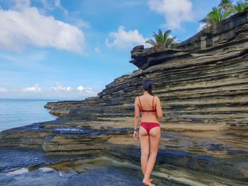 Rear view of bikini woman standing by rock formations at beach against cloudy sky