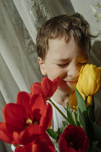 Toddler boy is holding lush bouquet of red and yellow tulips, making faces and smiling funny