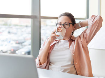Relaxed woman drinks coffee and works with laptop. modern office with panoramic windows