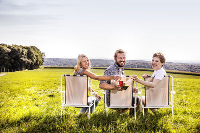 Happy friends sitting on camping chairs in rural landscape clinking bottles