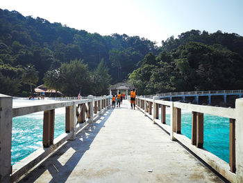 Rear view of people wearing life jackets while walking on footbridge over sea