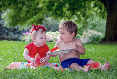 Young toddler boy shares watermelon with his baby sister as they sit outside on a summers day