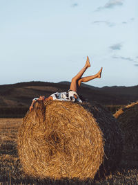 Blonde  woman in hat and white dress sitting on a haystack and enjoying the sunset. 