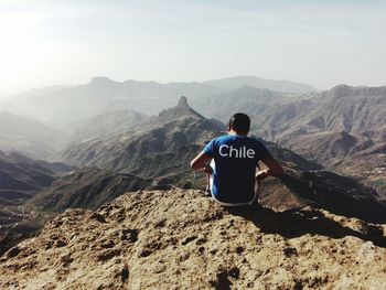 Rear view of man sitting on cliff against sky