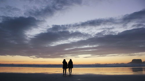 Silhouette friends standing on beach against sky during sunset