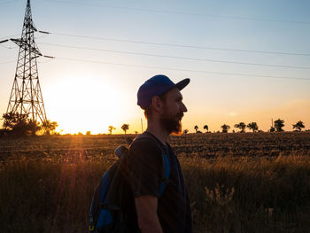 Side view of bearded man with backpack standing against sky during sunset countryside in the field