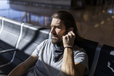 Adult bearded man in stylish outfit with hat sitting in a bench next to glass wall listening to music on wireless earphones while waiting for flight in airport lounge