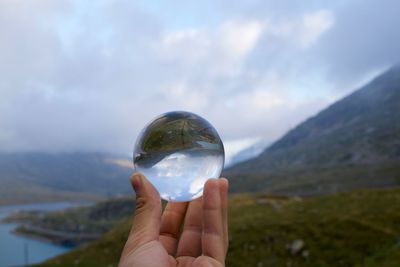 Cropped hand holding crystal ball over landscape against cloudy sky