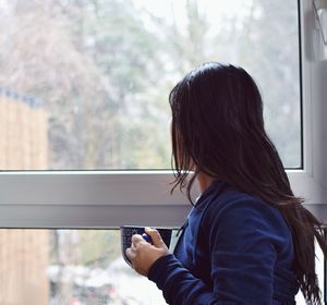 Side view of mid adult woman with drink looking through window at home