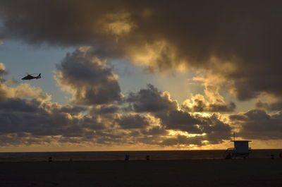 Silhouette helicopter flying over sea against cloudy sky during sunset